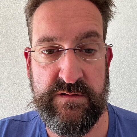 Portrait-Foto eines ca. 50jährigen Manner. Er hat weiße Haut, braune, kurze Haare und trägt einen Vollbart. Er sieht müde in die Kamera.